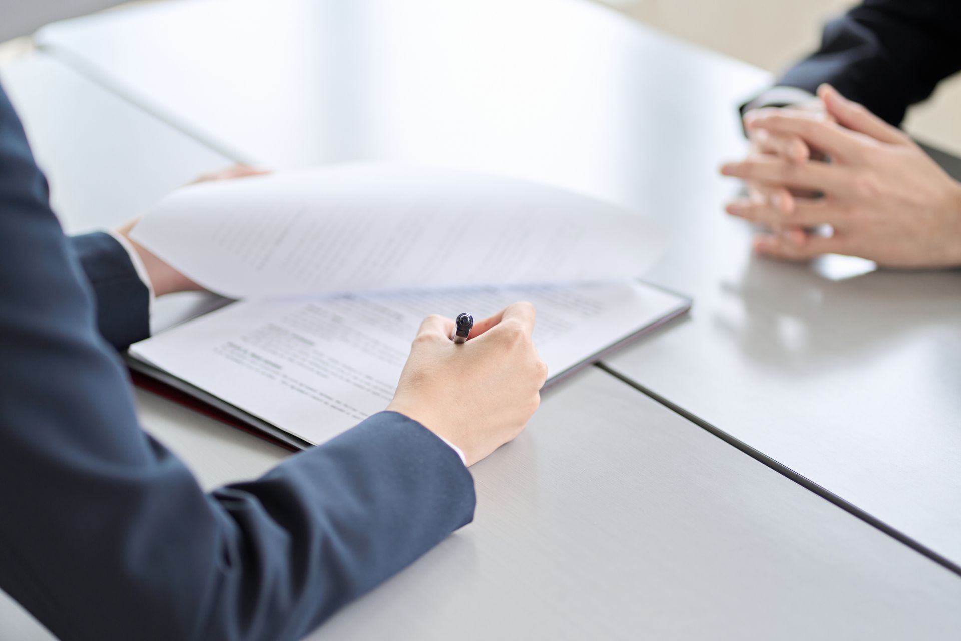 Person in a suit reviewing paperwork on a clipboard at a table with another person.