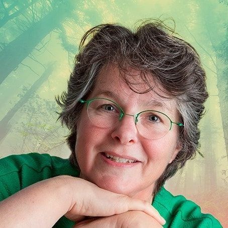 Woman with glasses smiles, resting chin on hands. Green shirt, curly gray hair. Forest background.