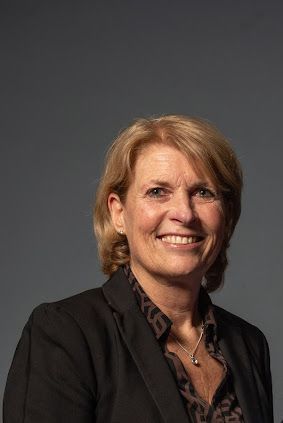 Woman with blonde hair wearing a black blazer and patterned shirt smiles against a grey backdrop.