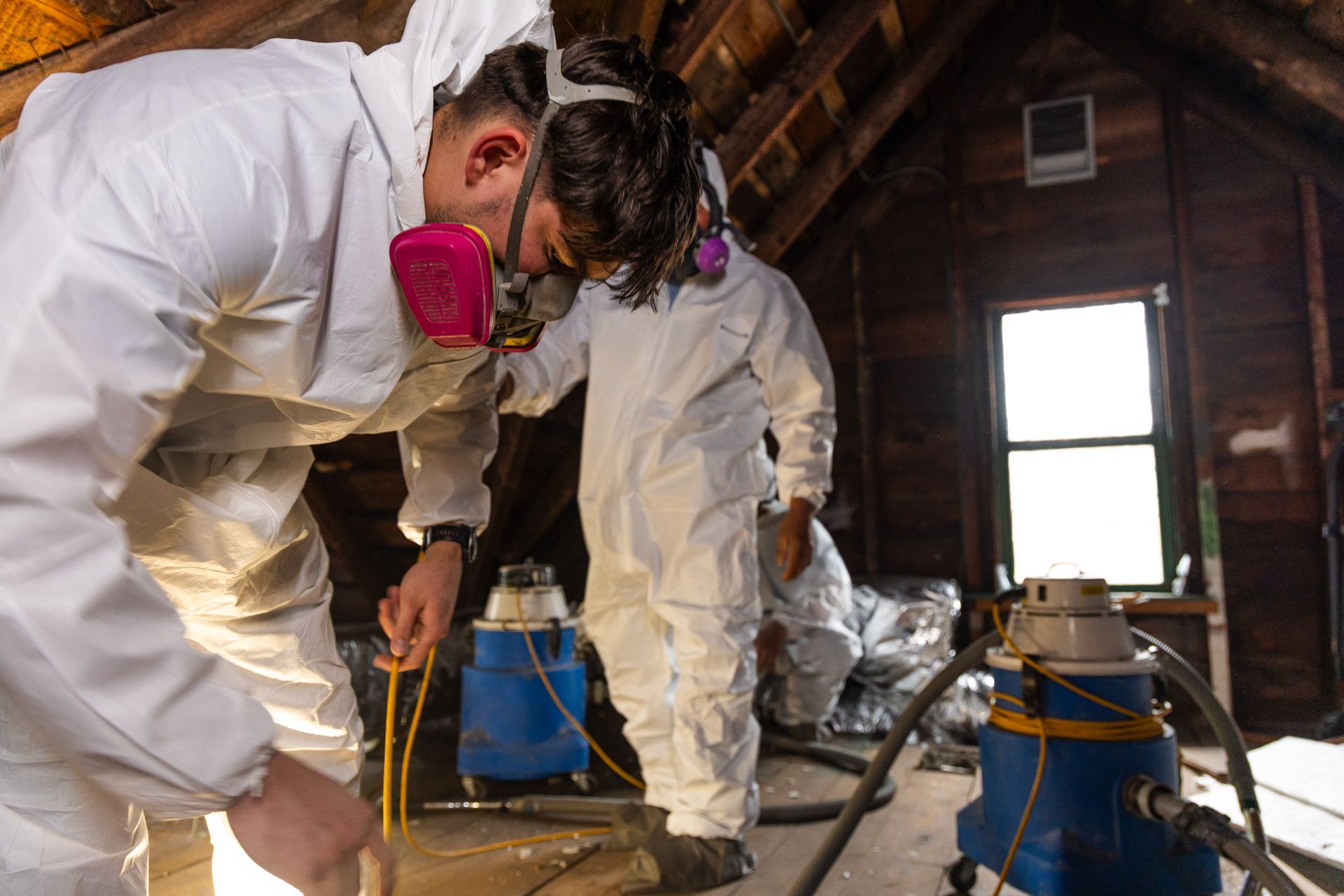 Workers in white suits and respirators inspect a dusty attic with equipment and tools