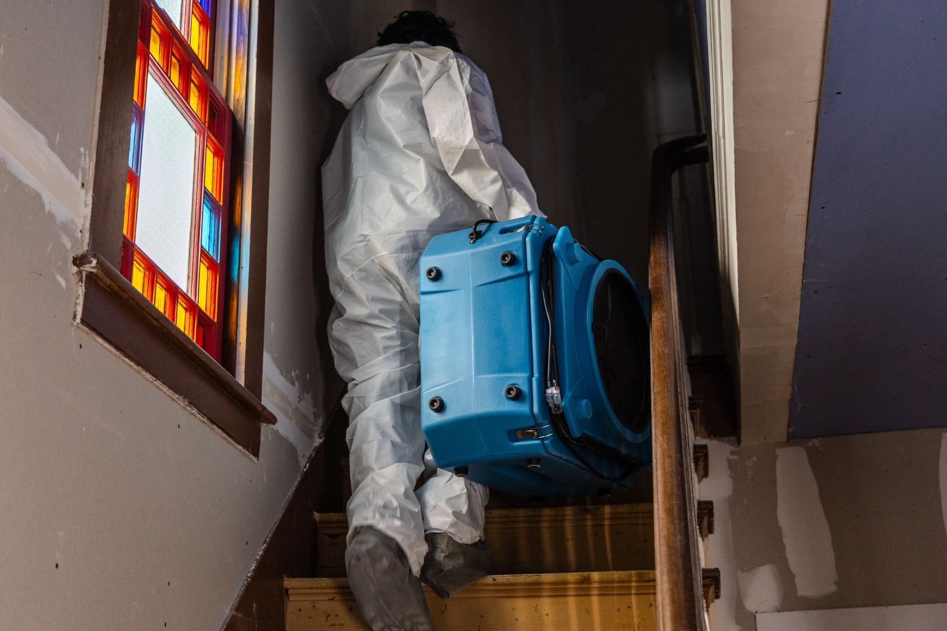 Person in white protective suit carrying a blue machine up a narrow staircase beside a stained-glass window
