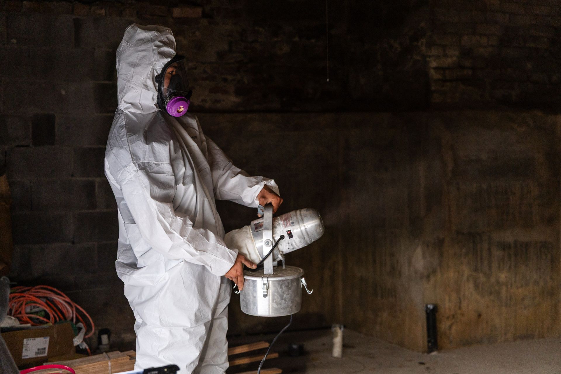 Person in a white hazmat suit pours liquid from a bucket in a dim industrial room.