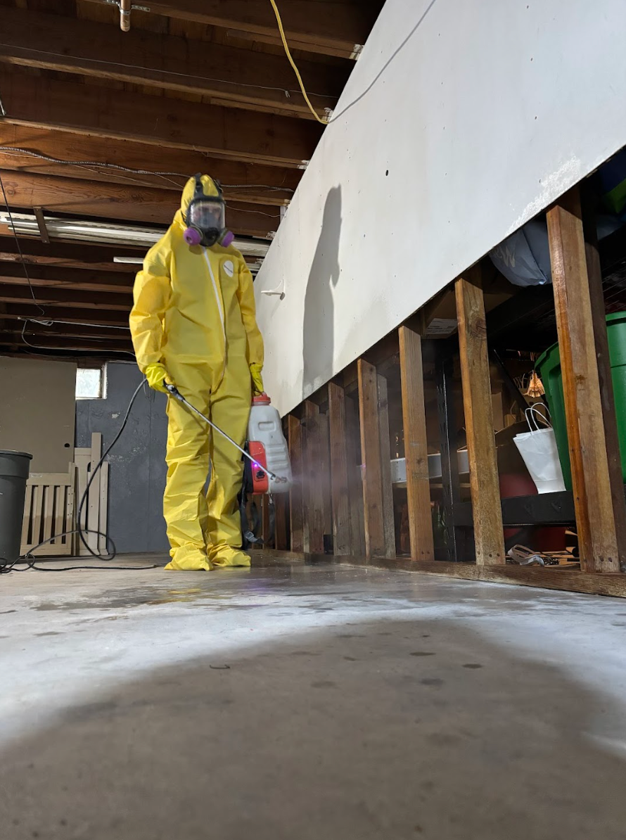 Person in yellow protective suit standing in a partially demolished room with exposed walls and debris.