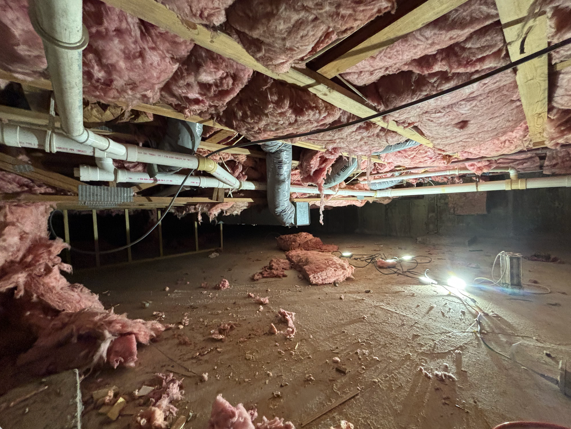 Crawl space with exposed pipes, pink insulation, and a dirt floor under a house