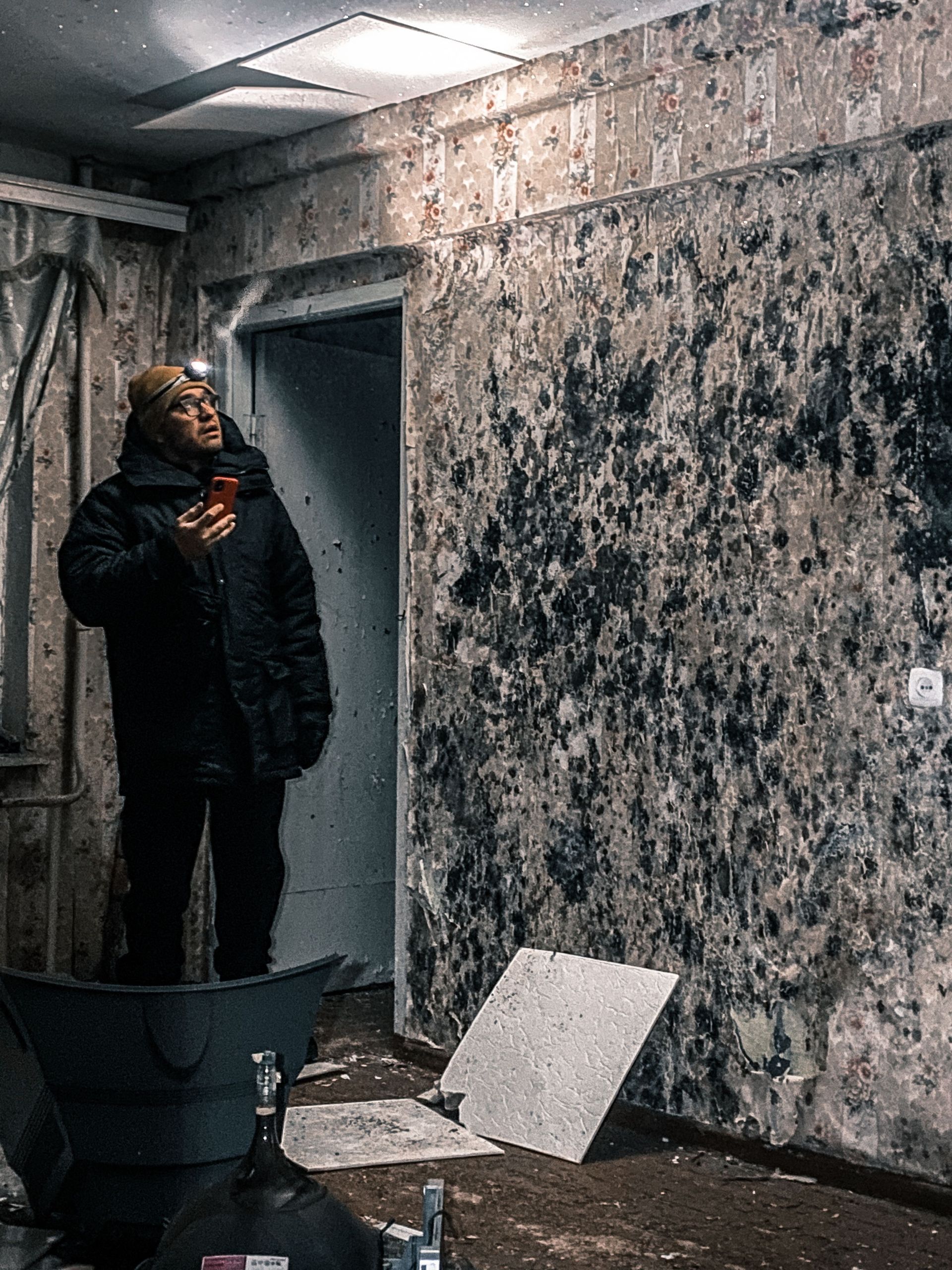 Person in a dark coat standing in a dim, worn room with peeling walls and scattered debris
