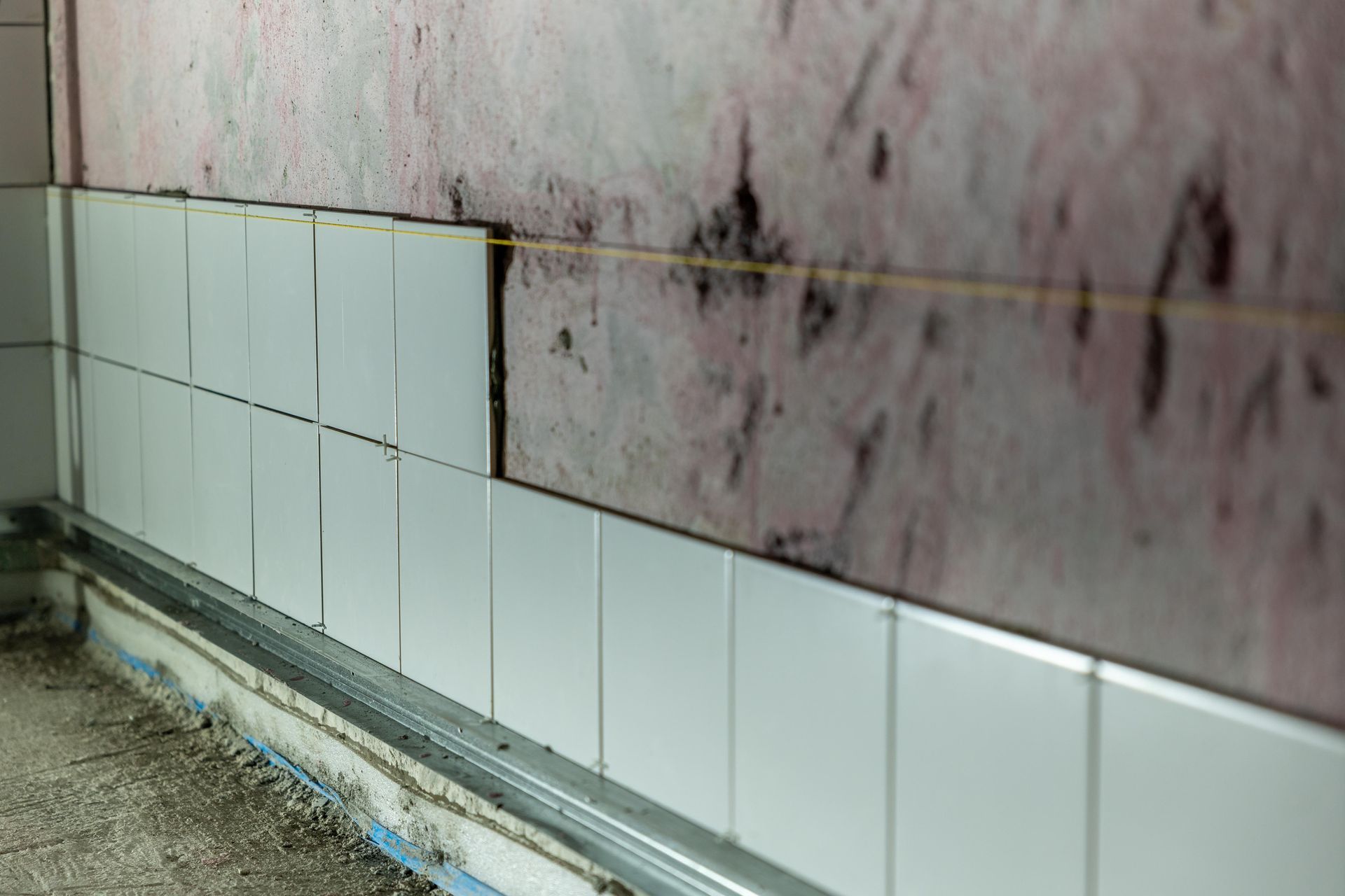 White wall tiles along a baseboard with damp, stained wall above and wet floor below.