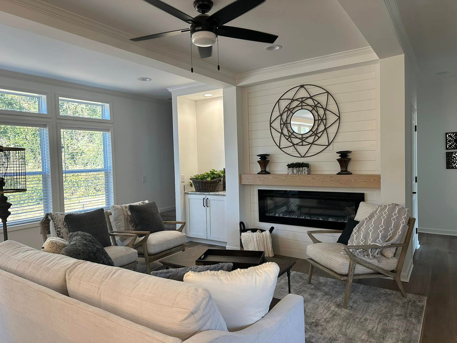 Bright living room with gray sofas, fireplace, ceiling fan, large windows, and circular wall decor