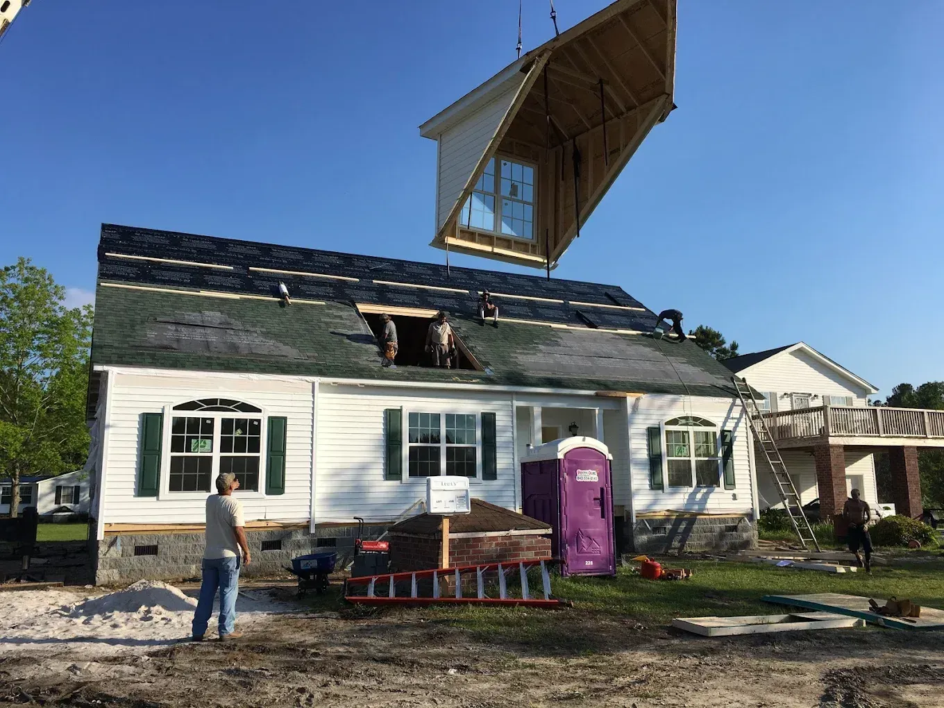 House with roof damage and a large structure being lifted above it, with people and a portable toilet outside