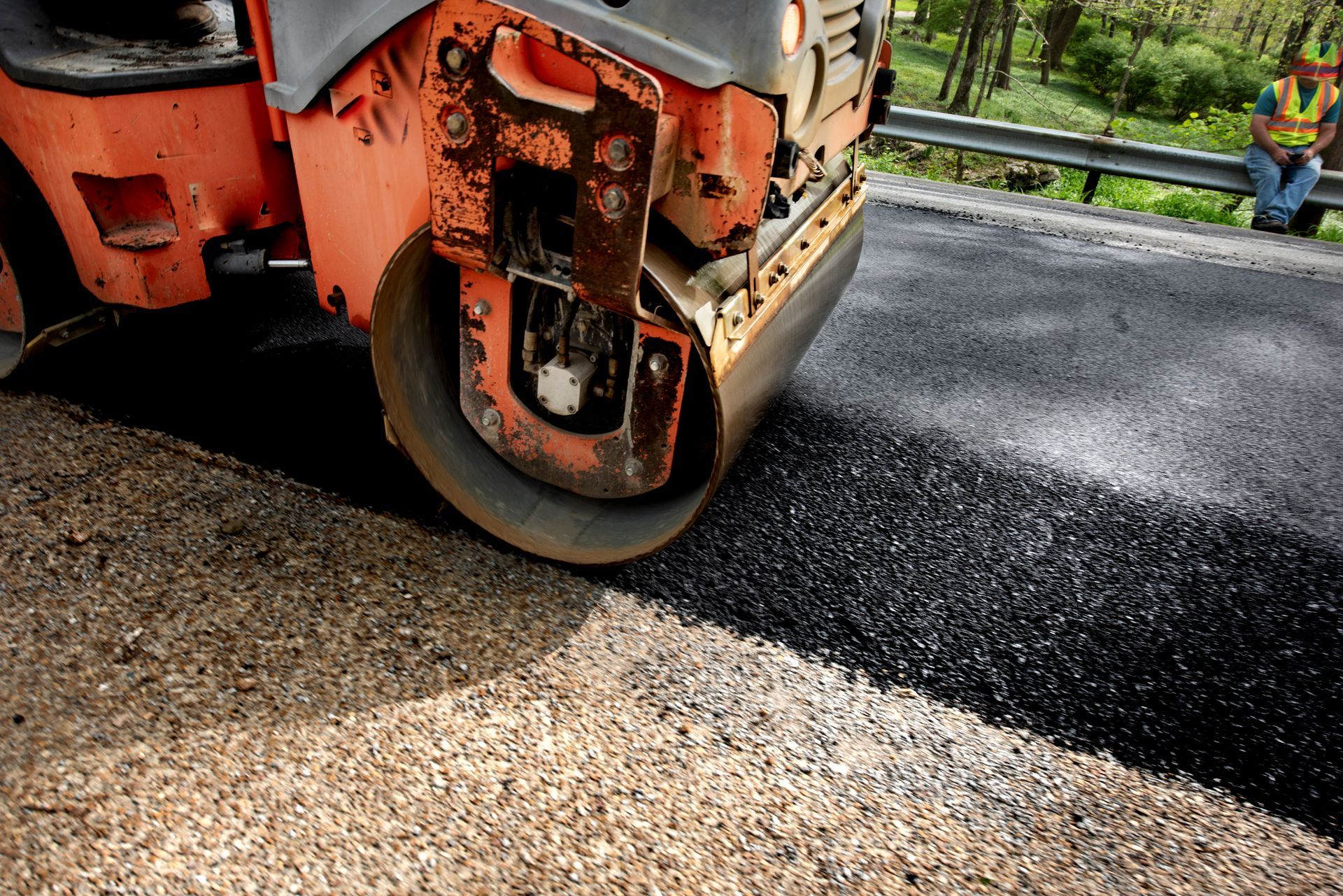 Road roller compacting fresh black asphalt on a road, with a worker in the background.
