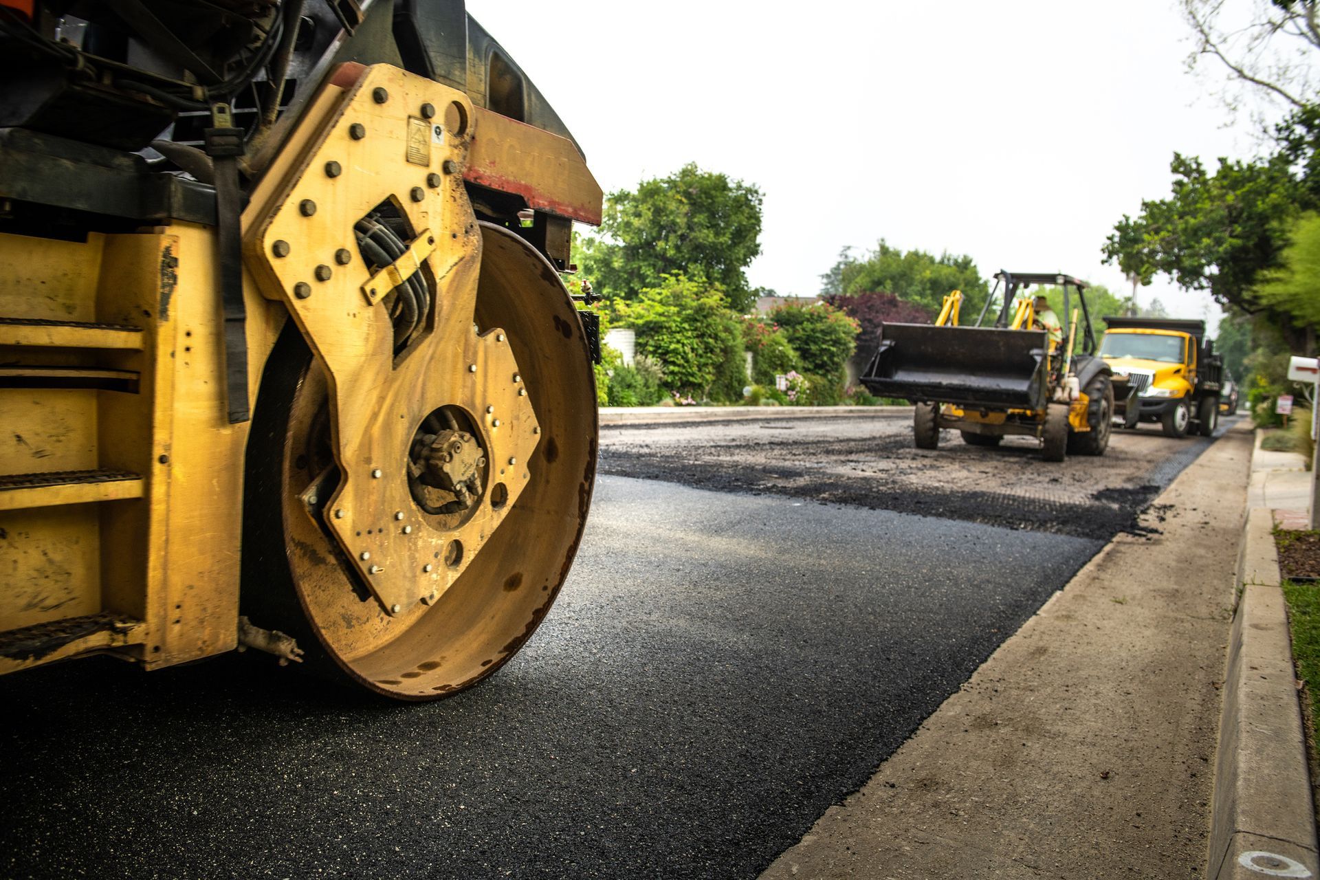 Yellow road roller compacting fresh asphalt on a residential street; construction equipment in background.