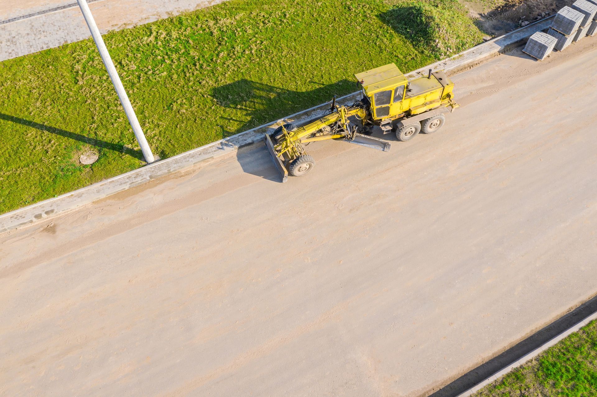 Yellow grader smoothing a dirt road. Beside a grassy hill and concrete blocks.