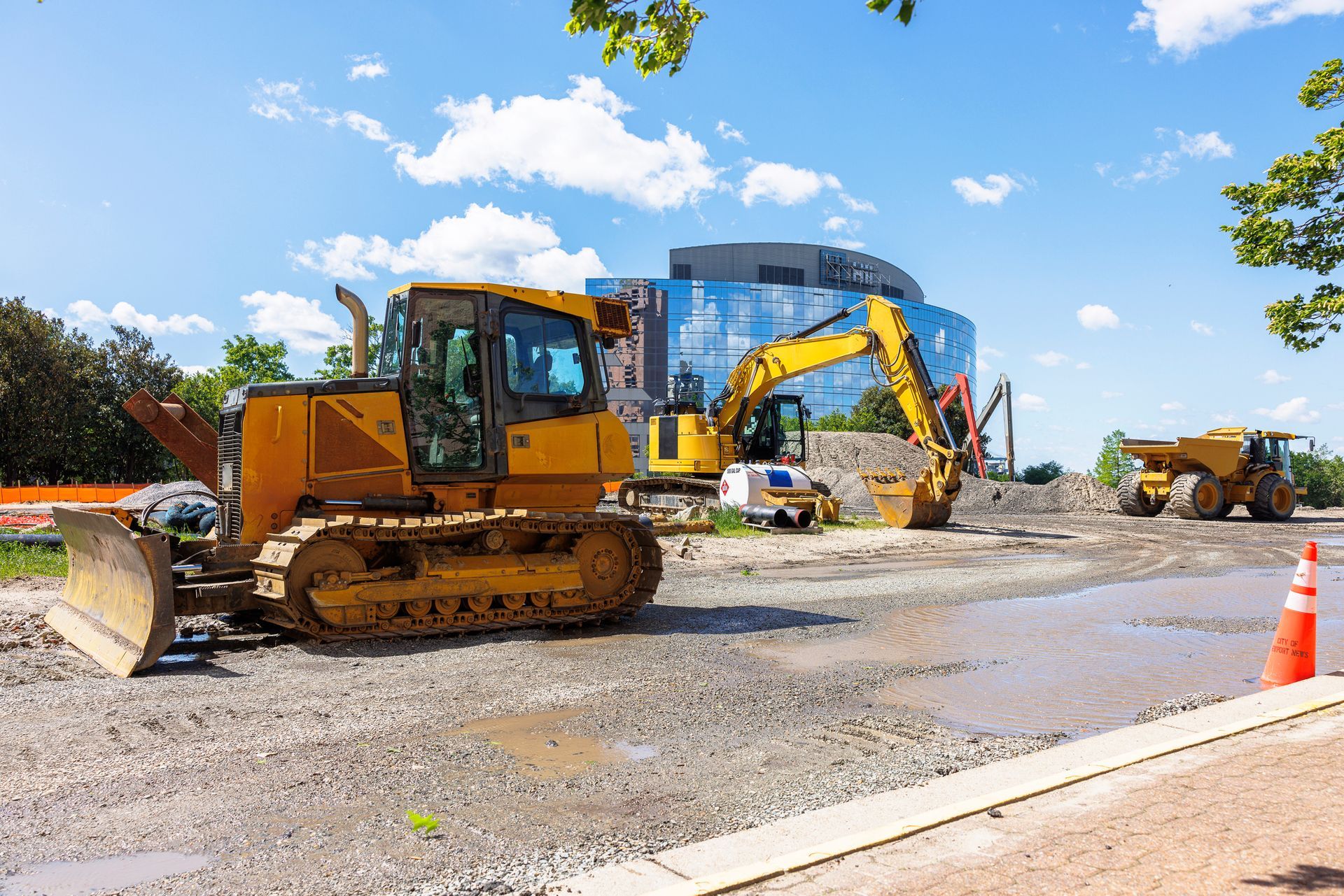 Construction site with bulldozer, excavator, and dump truck; blue glass building in background.