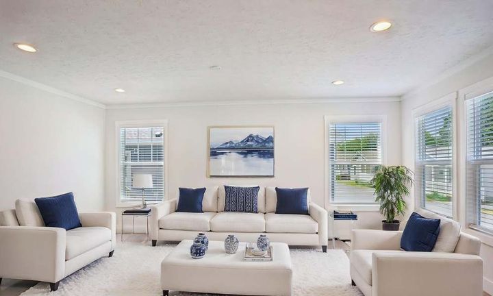 Bright living room with white furniture, navy pillows, and artwork. Windows allow natural light.