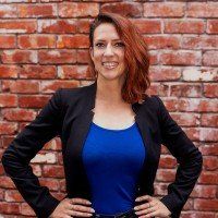 A woman in a blue shirt and black jacket is standing in front of a brick wall.