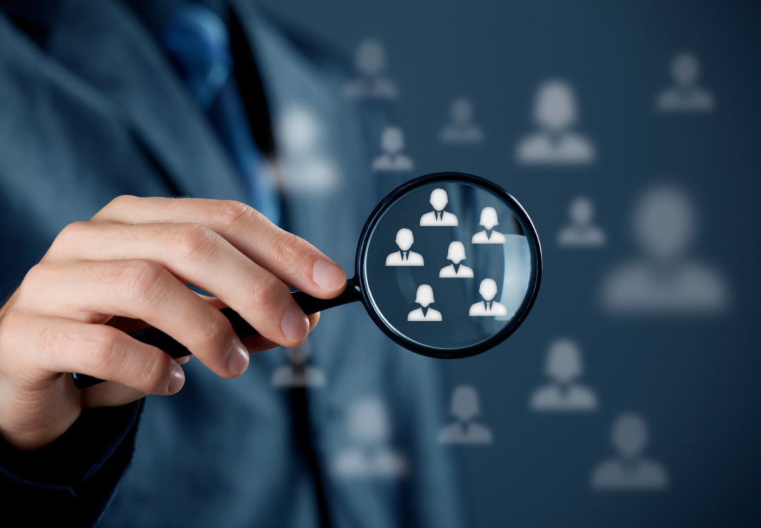 Person in suit holding magnifying glass over people icons, indicating focus on human resources.