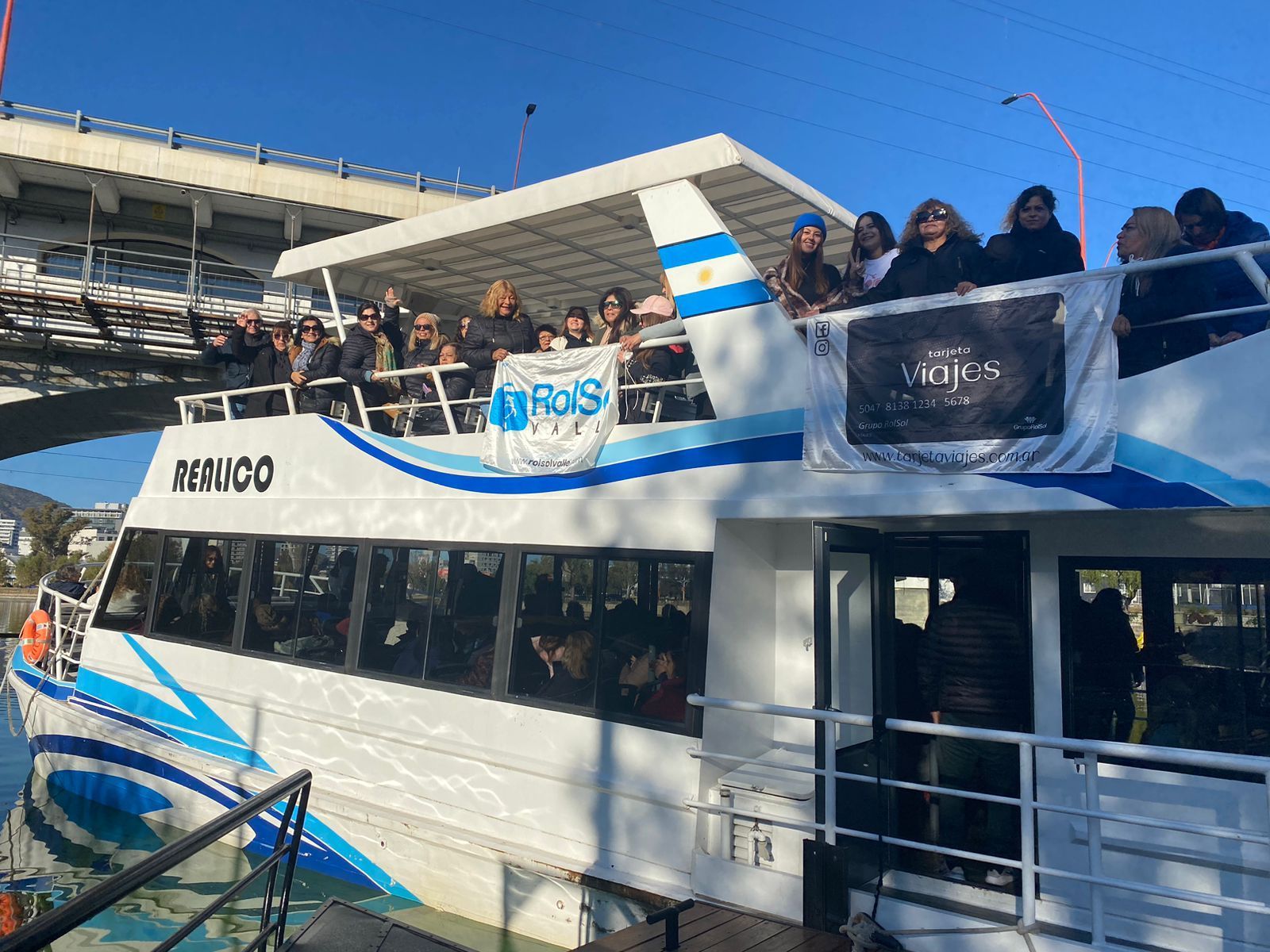 Personas en un barco con bandera argentina, pancartas y franjas azules y blancas; puente al fondo.