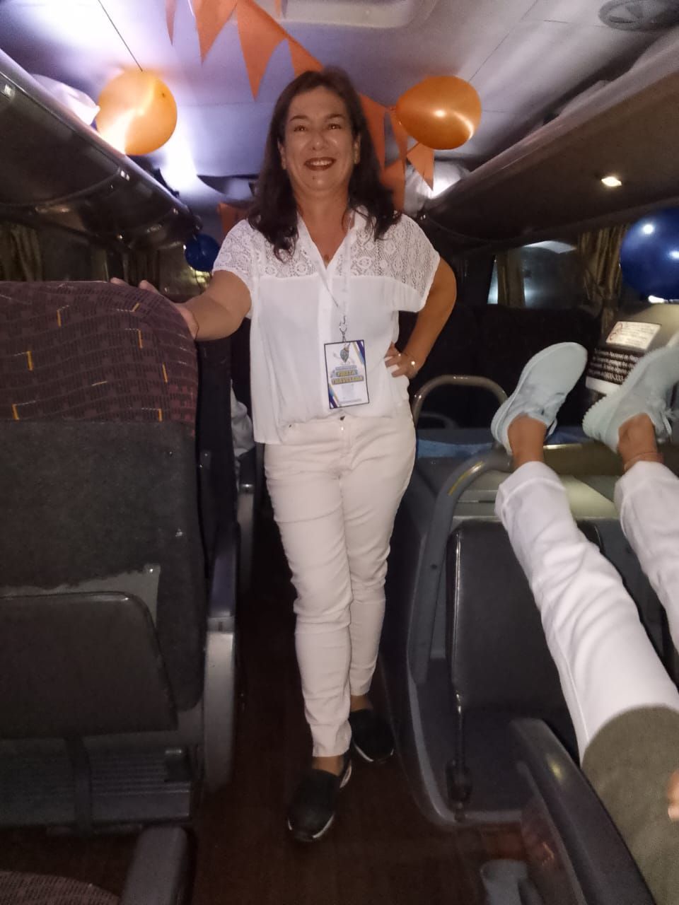 Mujer de pie en un autobús, sonriendo, vestida con camisa y pantalones blancos; globos naranjas sobre su cabeza.