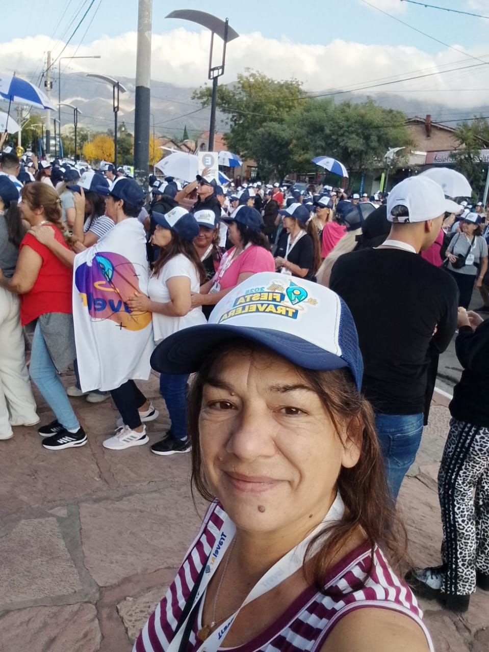 Una mujer se toma un selfi entre una multitud con sombreros blancos y azules. Al aire libre, un día soleado.