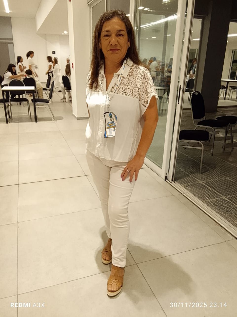 Una mujer vestida de blanco se encuentra en un interior moderno y bien iluminado, cerca de puertas de vidrio.