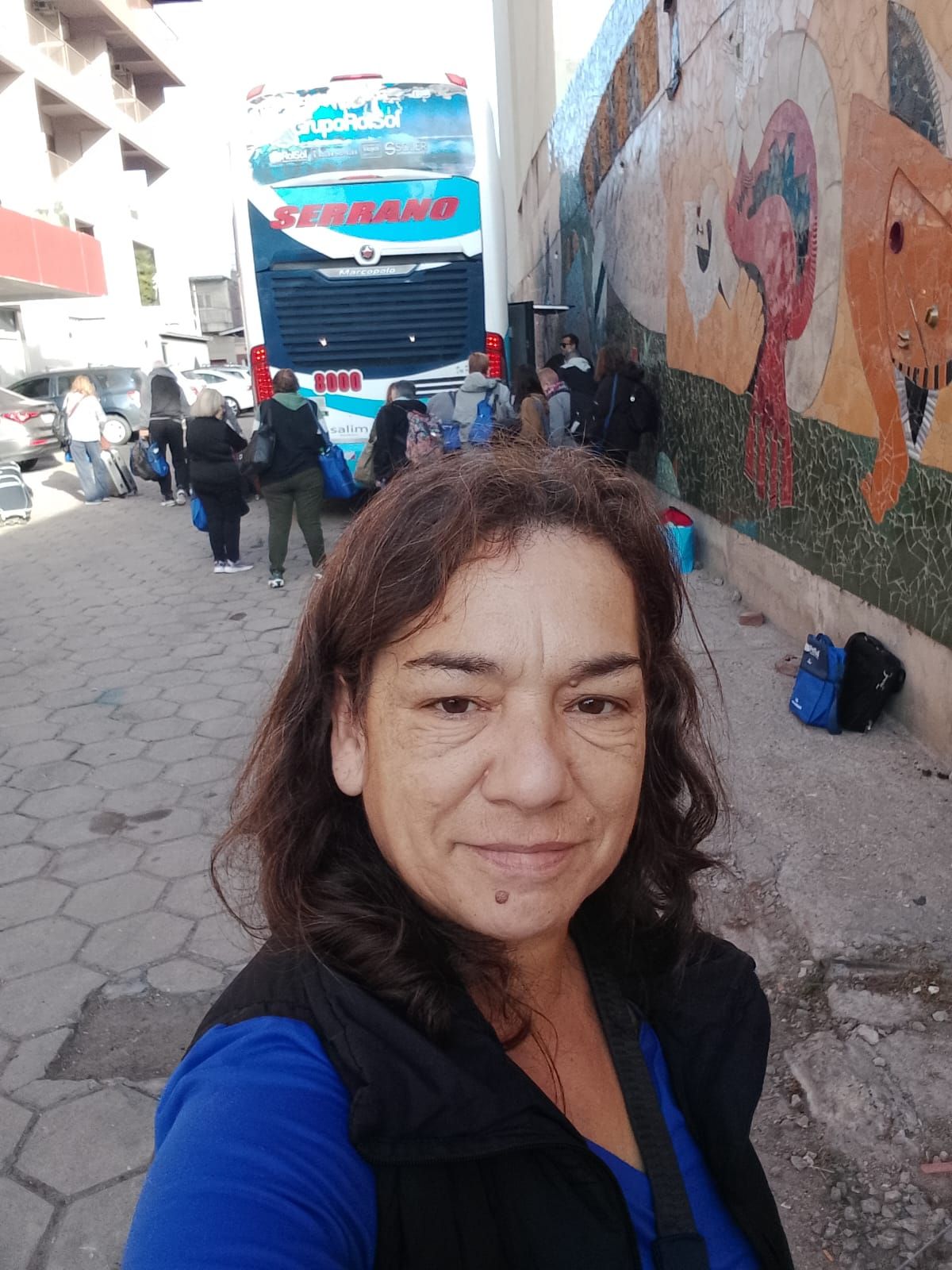 Una mujer sonríe a la cámara, pasajeros cerca de un autobús con una pared mural al fondo.