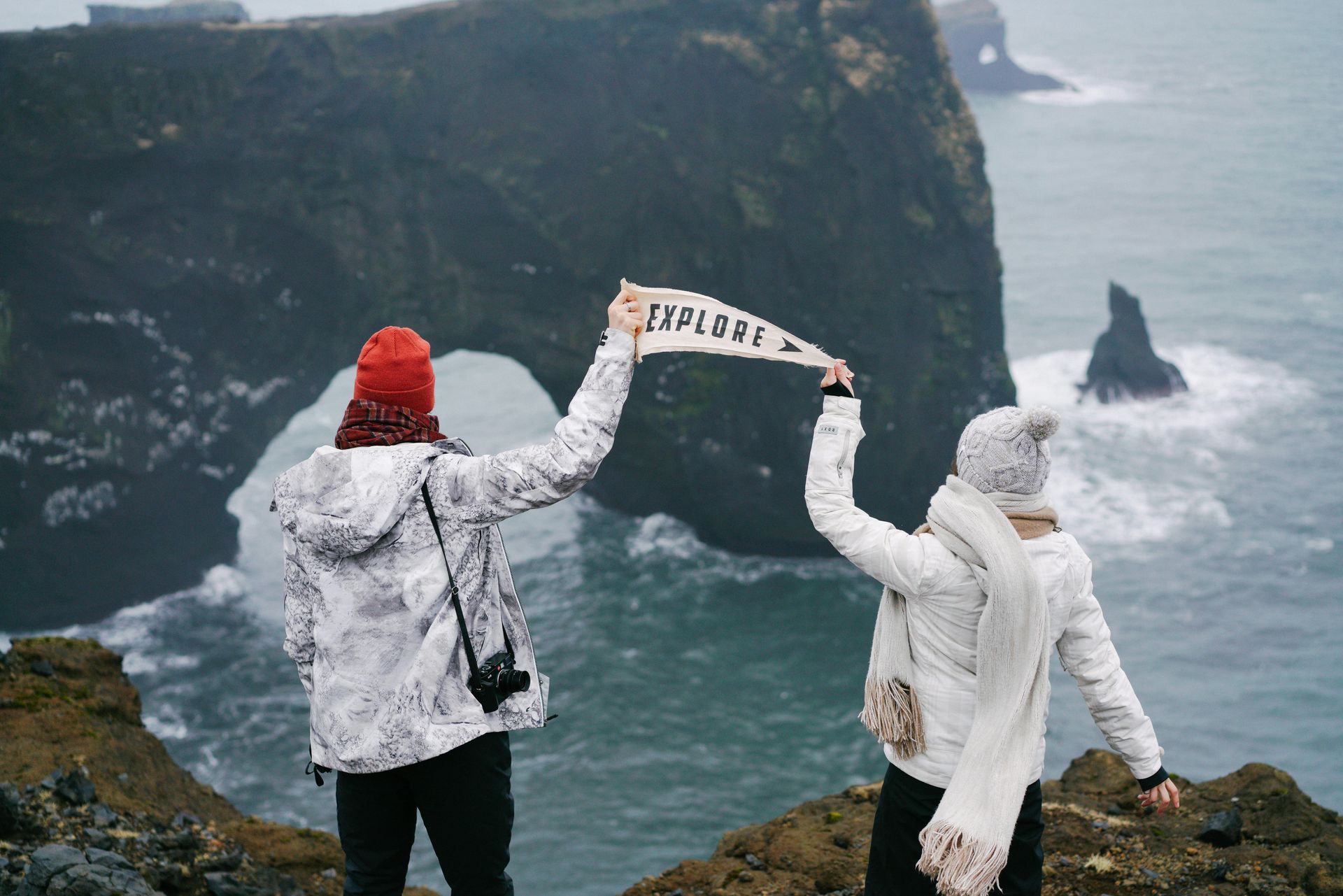 Dos personas se encuentran al borde de un acantilado, con vistas a un arco rocoso junto al mar, sosteniendo un banderín 