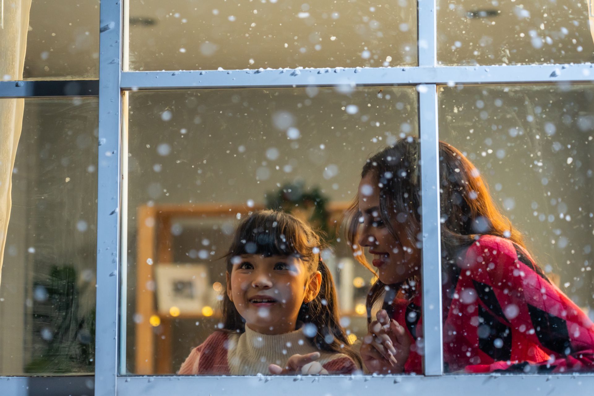 A woman and a little girl are looking out of a window at the snow.