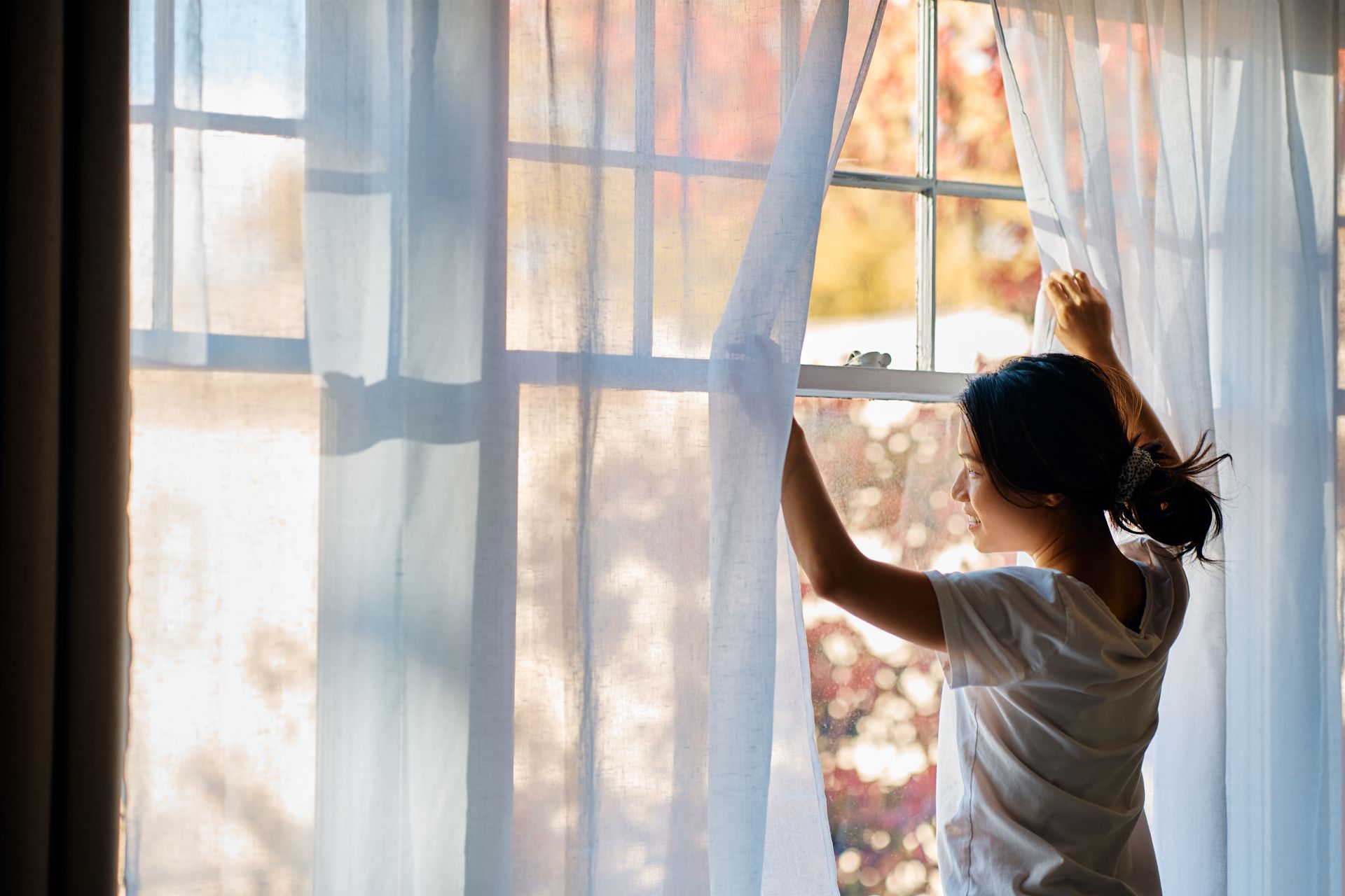 A woman is opening her curtains and looking out of a window.