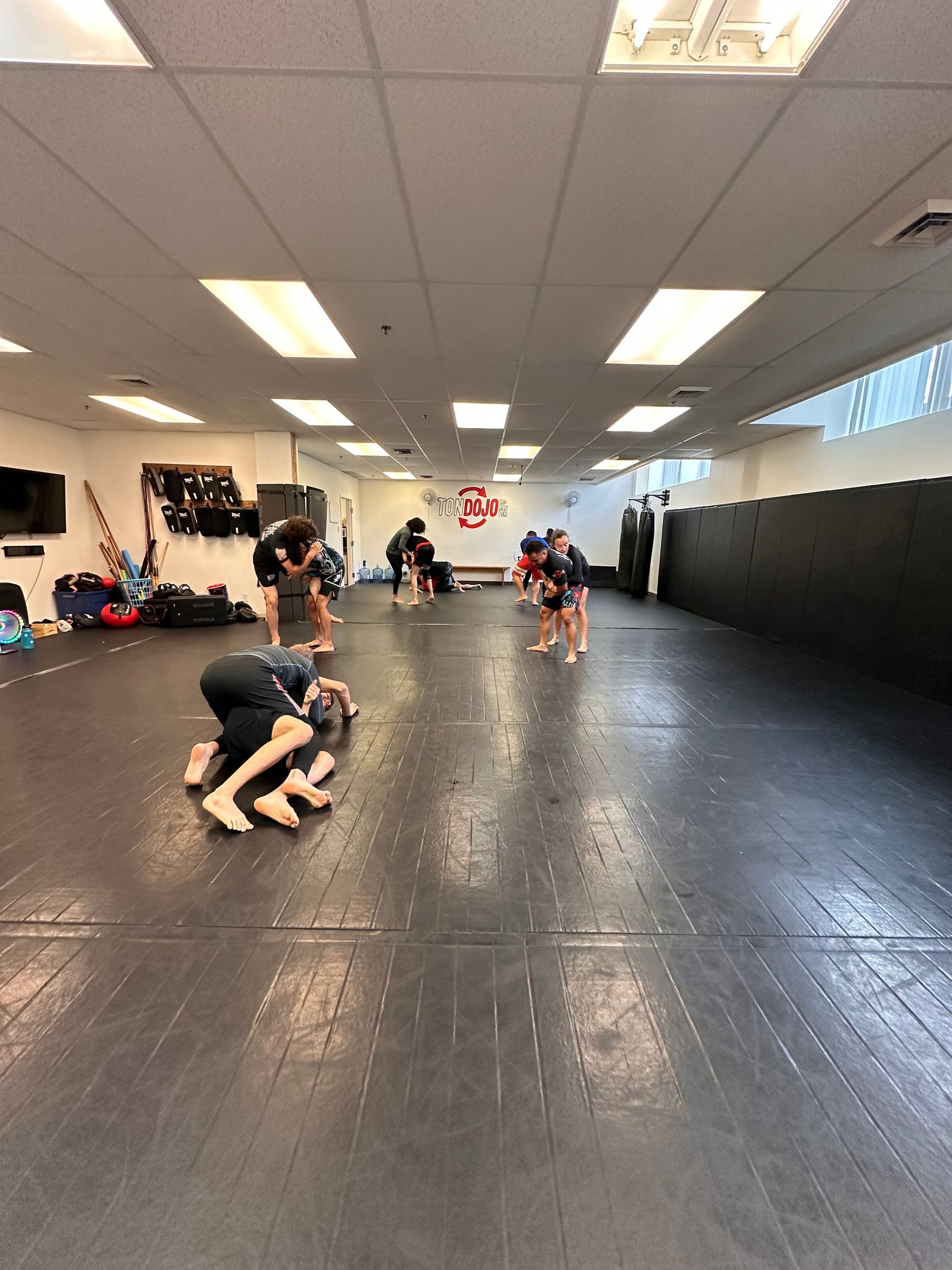 Des gens s'entraînant au grappling sur un grand tapis noir dans une salle de sport bien éclairée.
