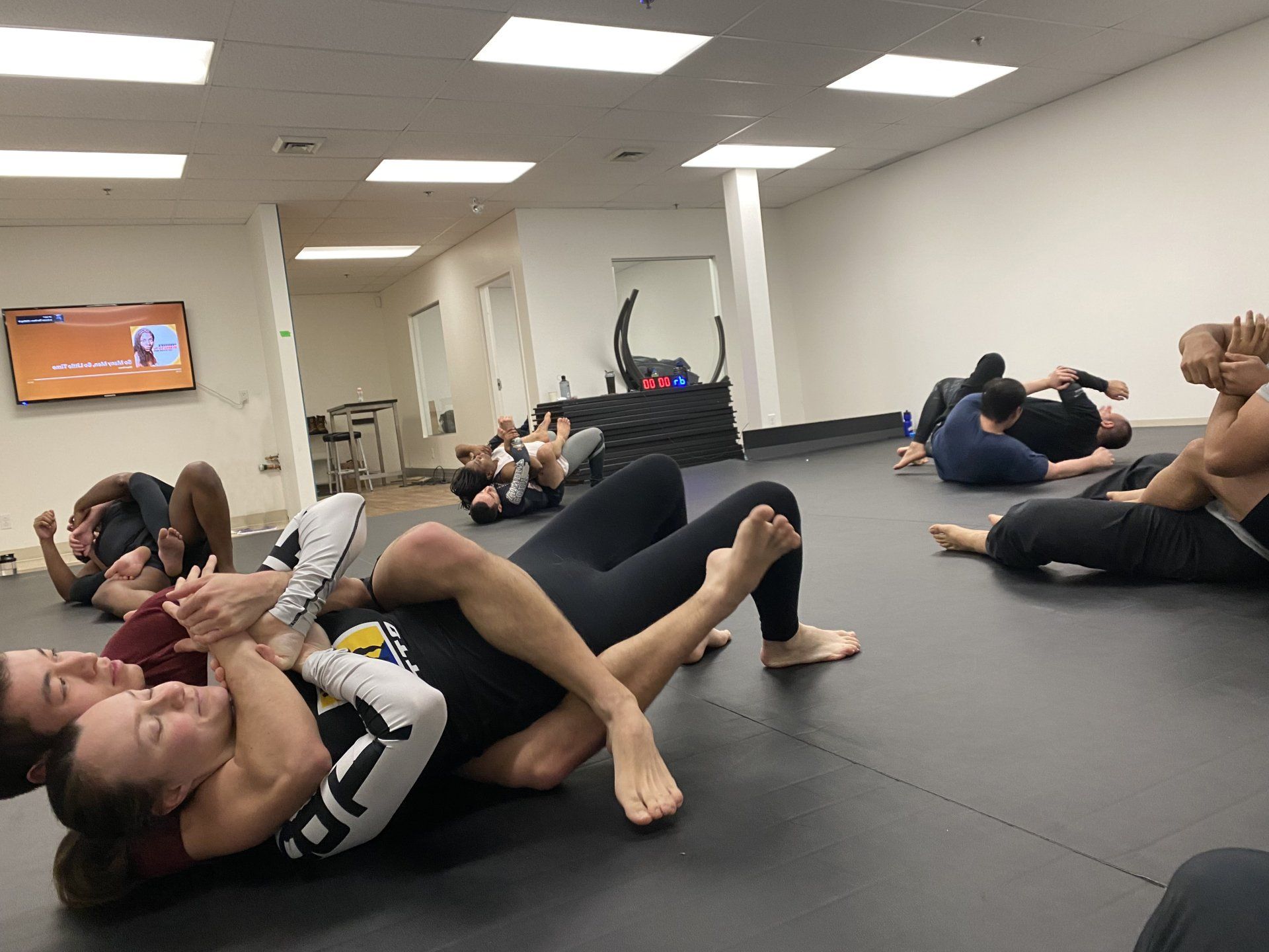 Des pratiquants de Jiu-Jitsu sur un tapis noir dans une salle de sport. Certains luttent, d'autres sont assis.