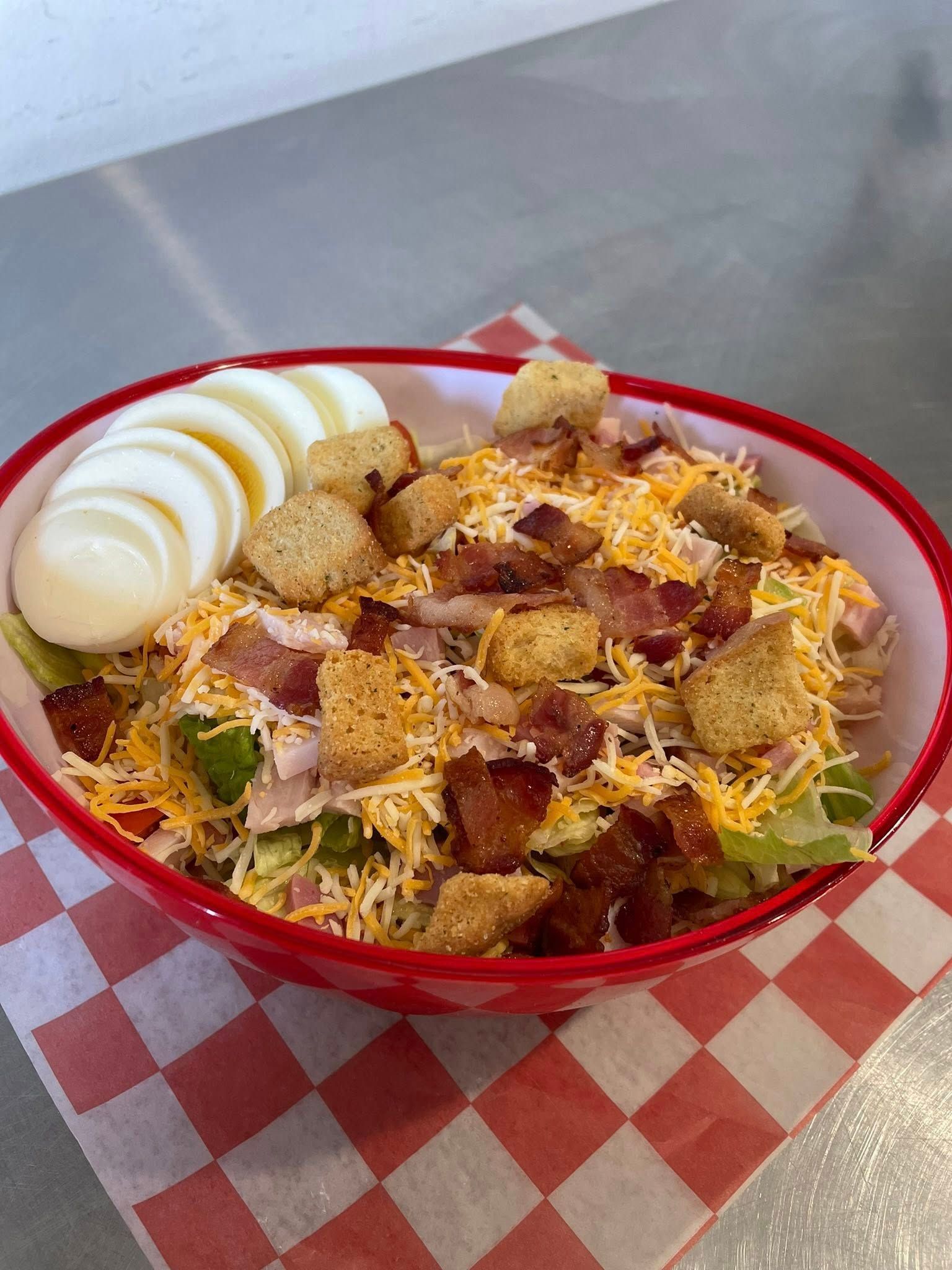 Chicken and bacon salad with hard-boiled eggs, croutons, and shredded cheese in a red bowl on a checkered napkin.