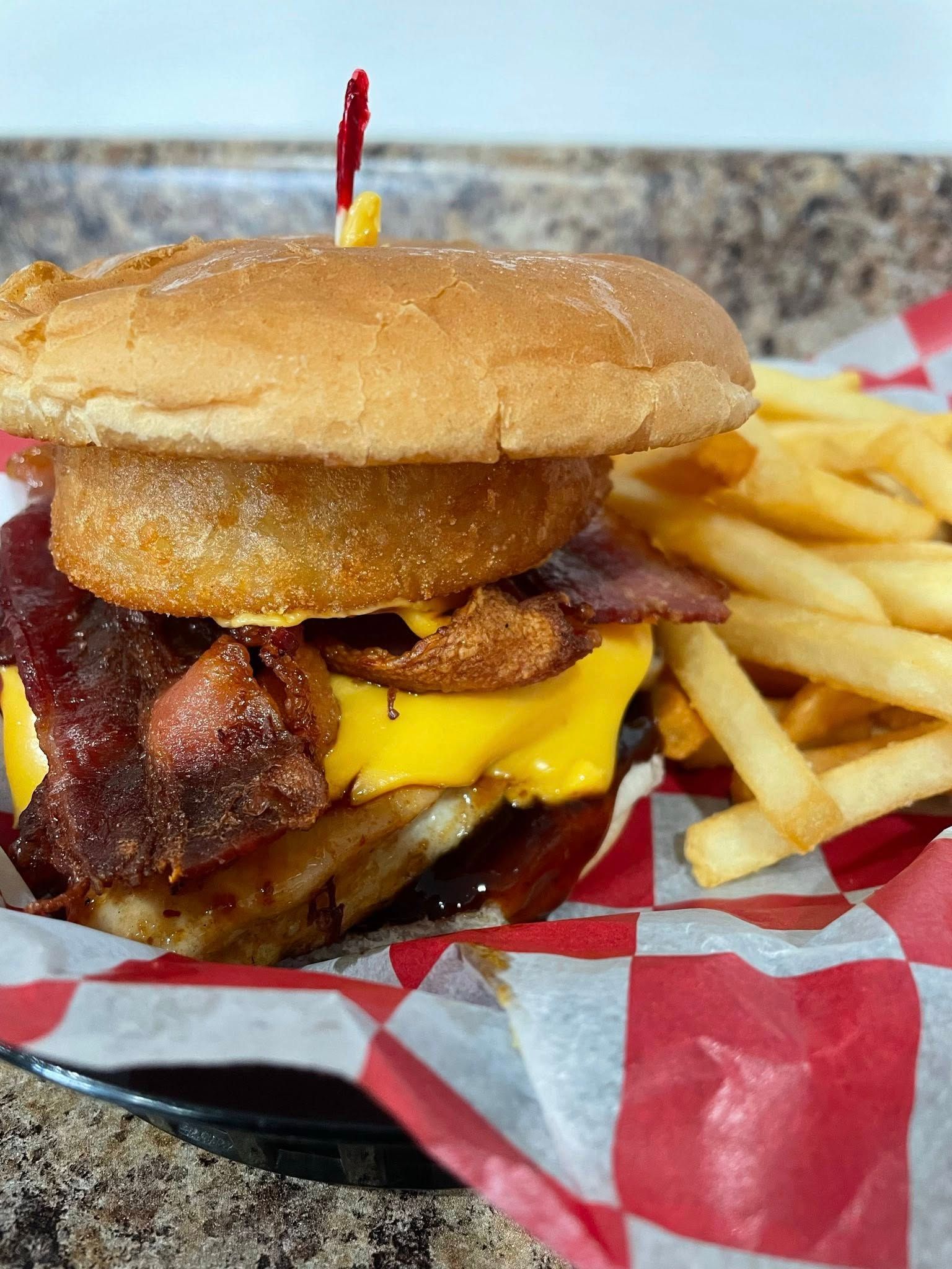 Burger with cheese, bacon, and onion ring, served with fries in a red and white checkered basket.