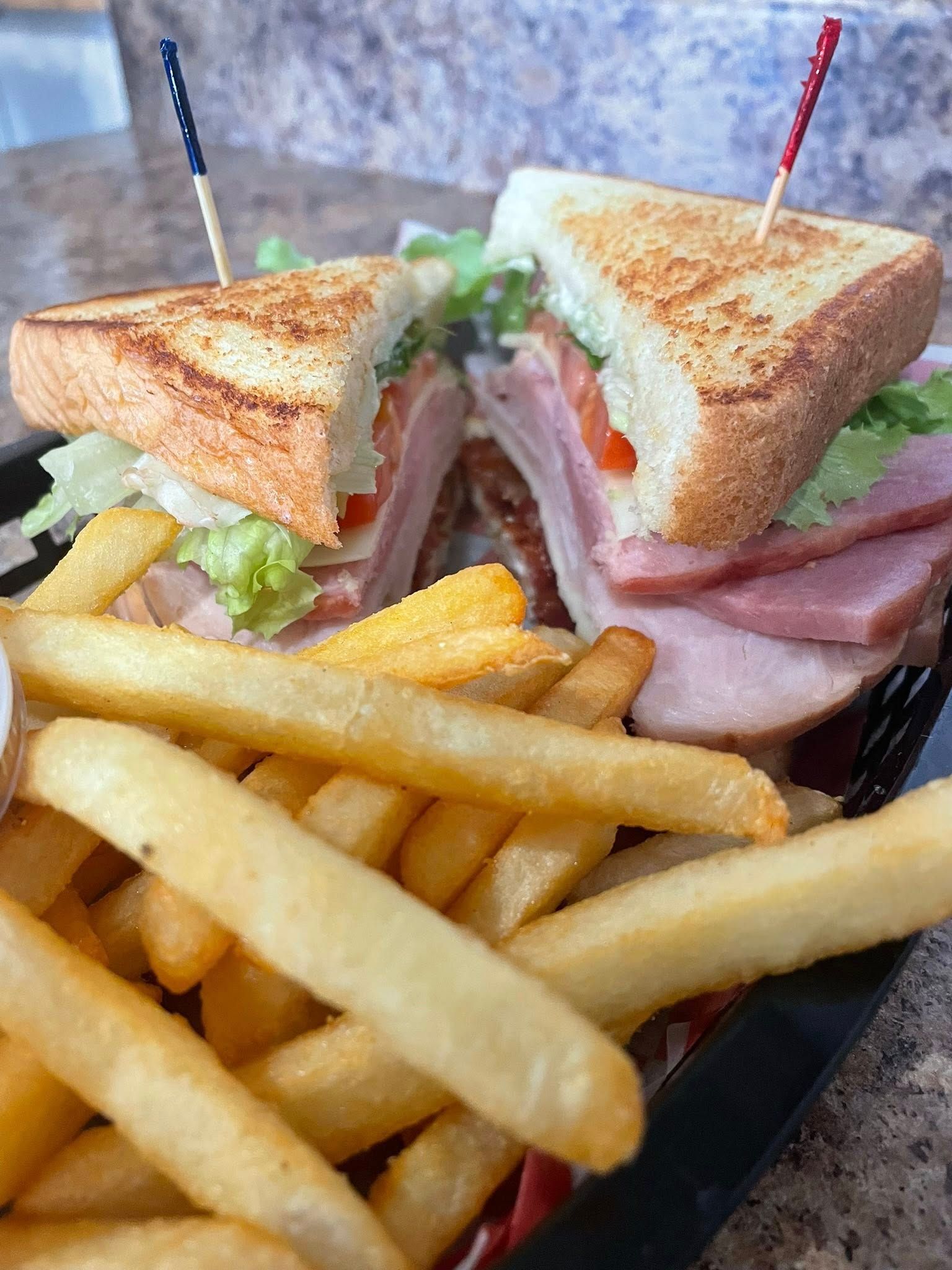 Club sandwich with ham, bacon, and lettuce, cut in half, served with fries in a basket. Toothpicks hold sandwich together.