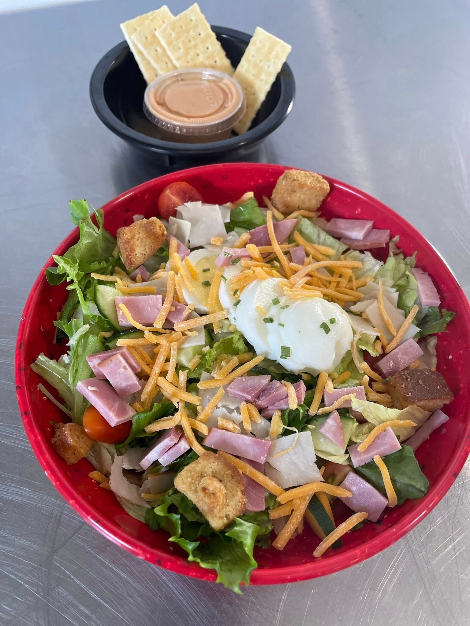 Salad with ham, cheese, croutons, and a poached egg in a red bowl, served with crackers and dressing in a small black bowl.