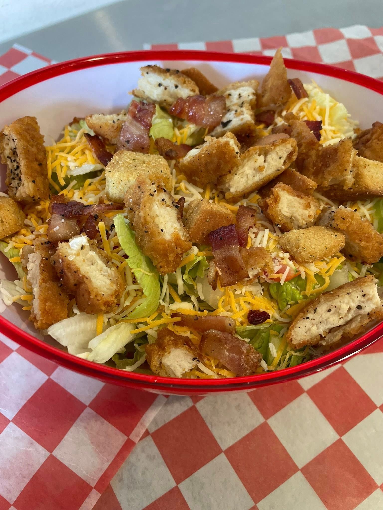 Salad bowl with fried chicken pieces, bacon, shredded cheese, and lettuce, set on a red checkered surface.