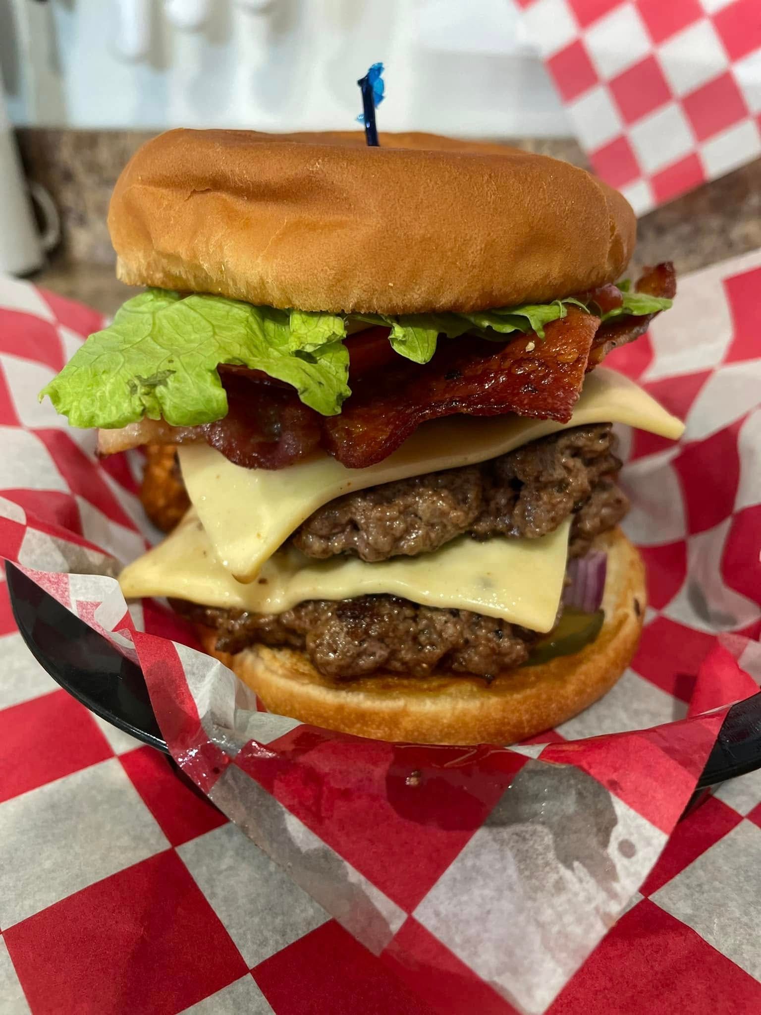 A hamburger with cheese , bacon , lettuce and pickles is sitting on a checkered napkin.