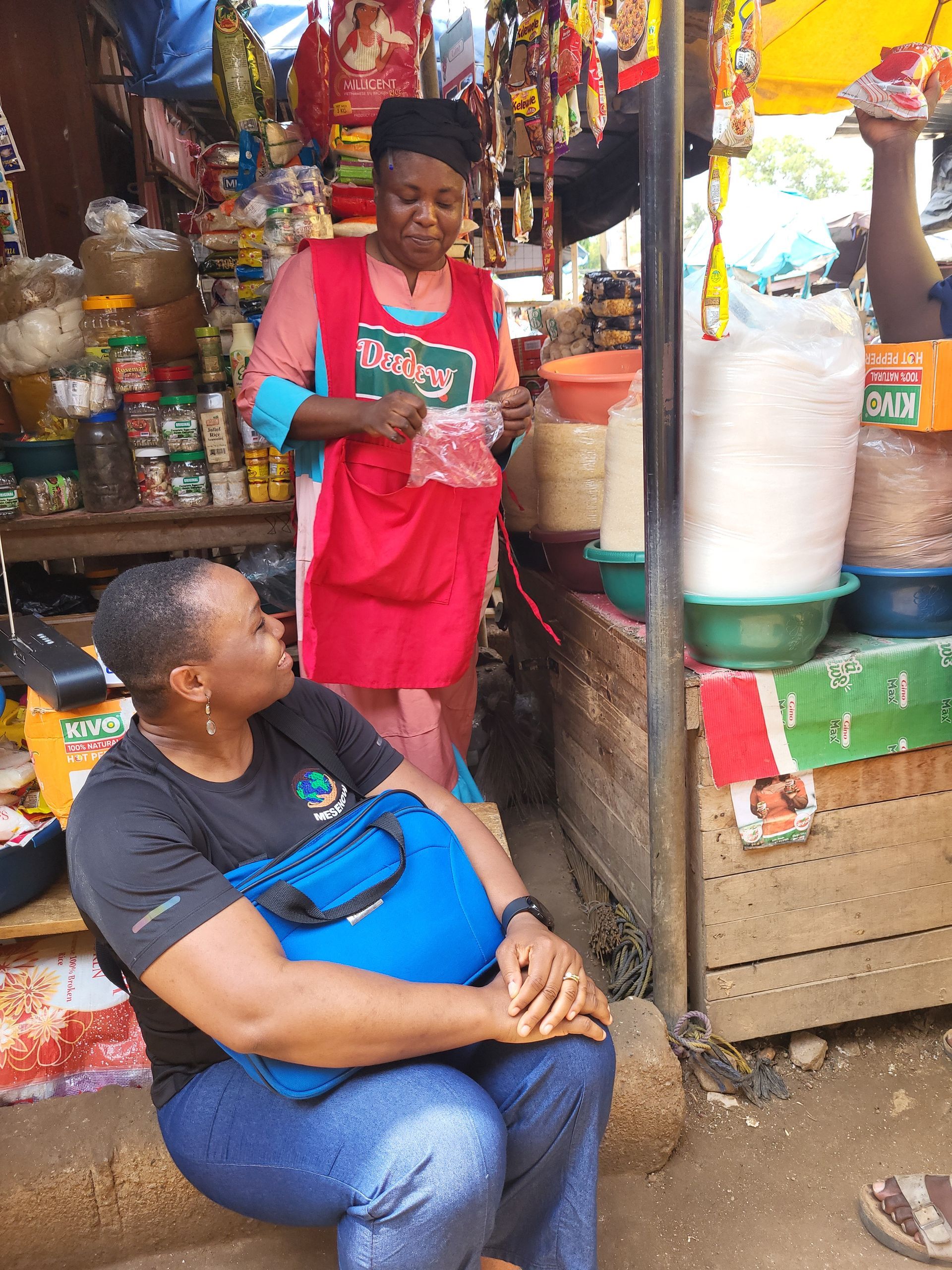 Woman in blue jeans and black shirt talking to a vendor wearing a red apron at a market stall.