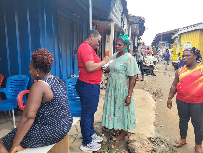 Man in red shirt talks with woman in green dress in a crowded outdoor market. Other people nearby.