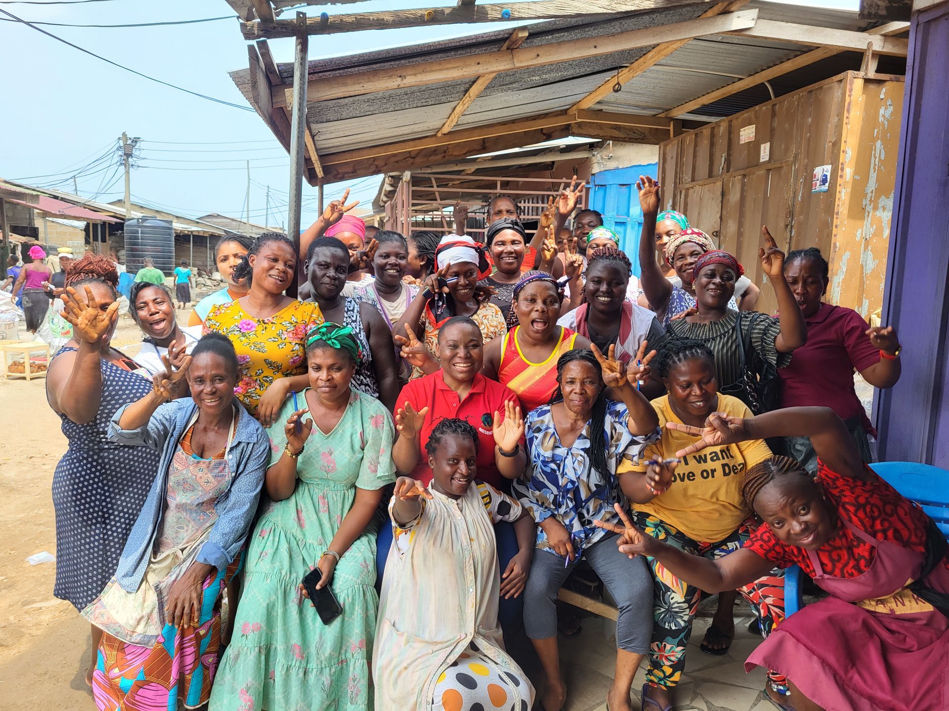Group of women smiling and waving in front of market stalls. Many are in colorful dresses.