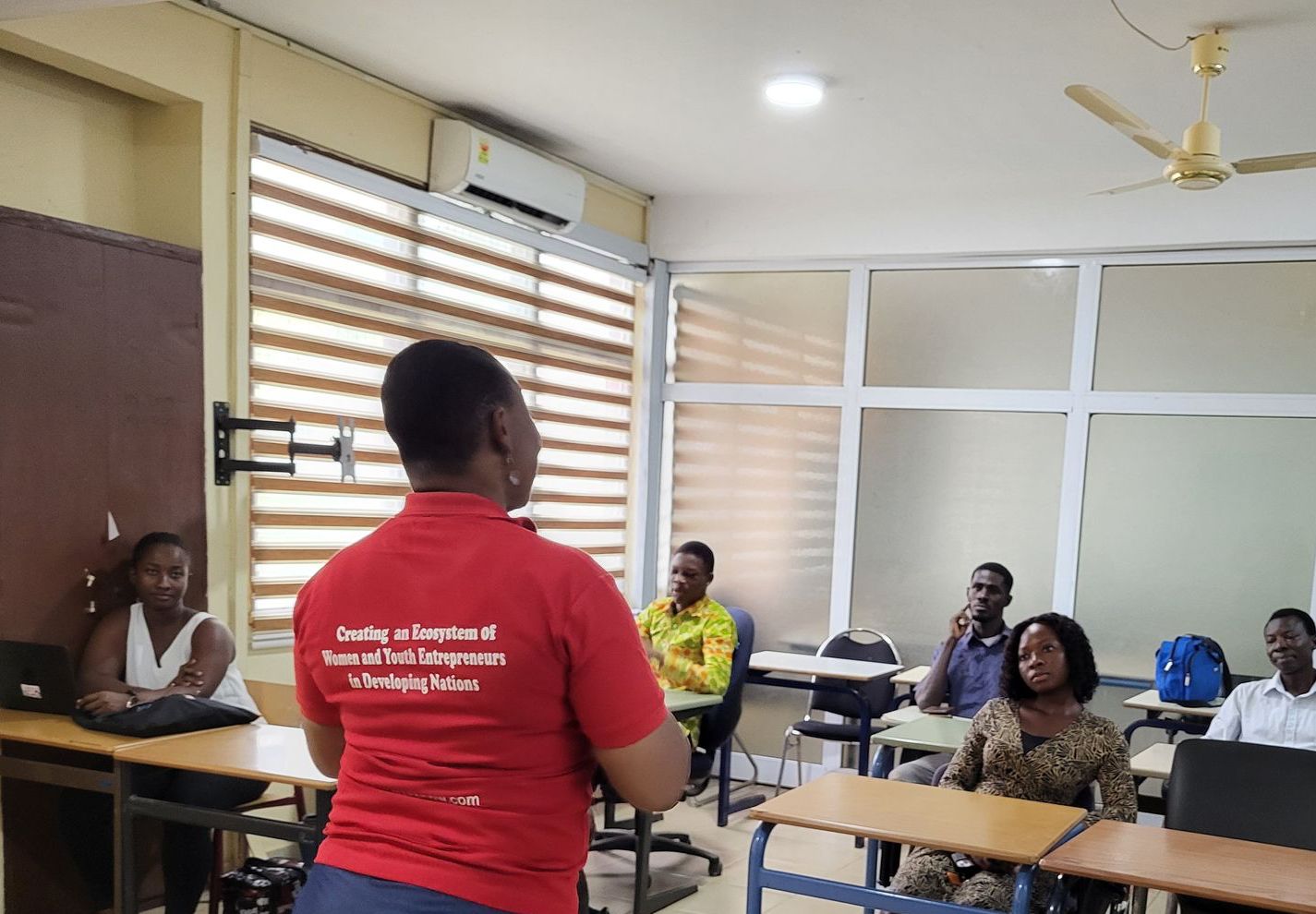 Woman in red shirt addresses a group in a classroom. Students sit at desks, some with laptops.