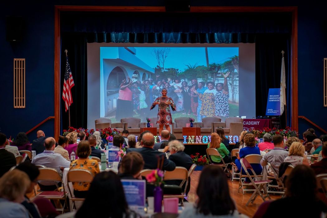 Audience watches presentation on screen at event; speaker stands on stage.