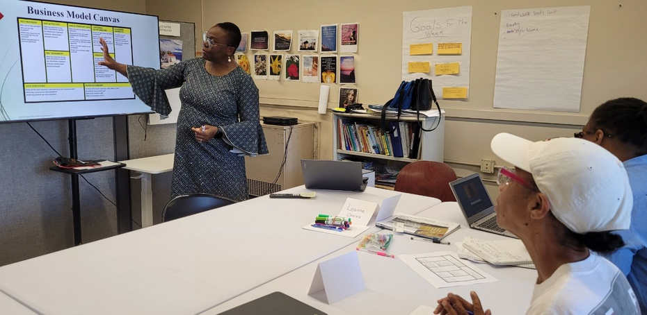 A woman points at a screen during a presentation. Several people sit at a table, watching.