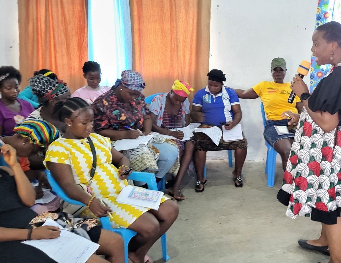 Group of women in a workshop, listening to a speaker. Some are taking notes. Bright interior, some wear colorful clothes.