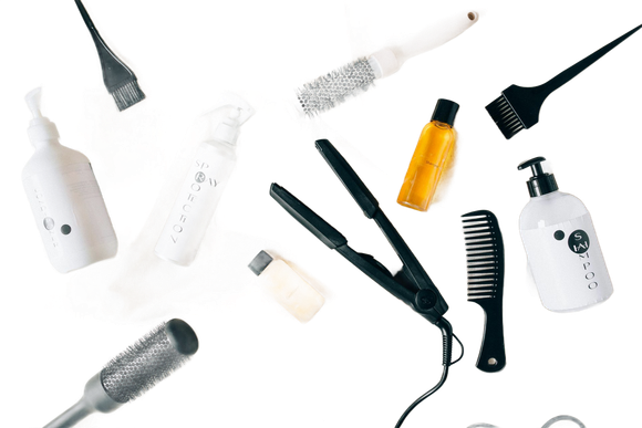 Hair styling tools scattered on white background: flat iron, brushes, bottles.