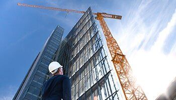 Engineer Working On Construction Site With Laptop — Steel Reinforced Concrete in Fort Worth, TX Construction worker in hardhat looking up at a tall building under construction with a crane, sunny sky.