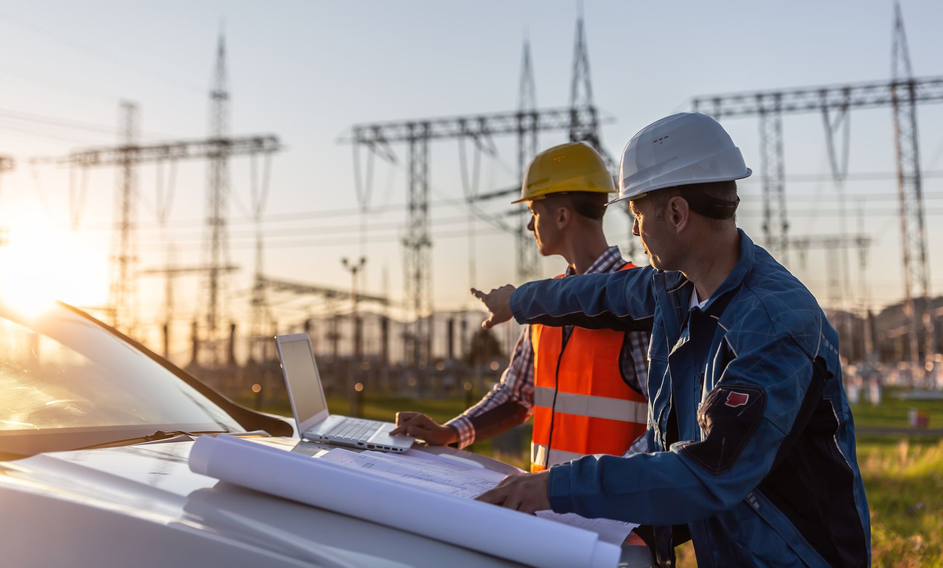 Structural engineers in safety helmets reviewing blueprints and using a laptop at a power substation site