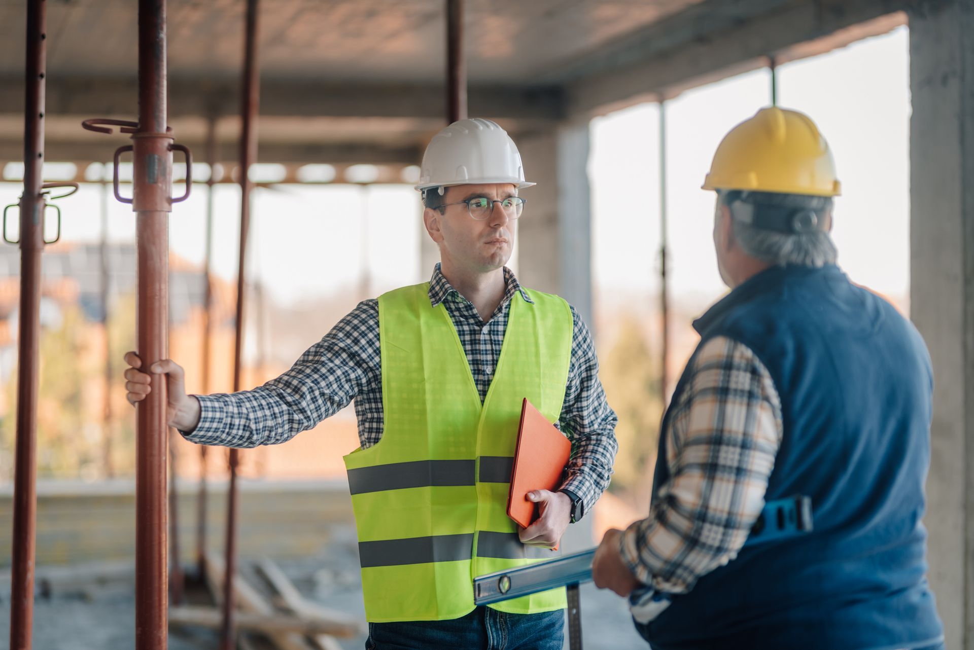Two engineers wearing safety vests and hardhats are discussing building progress at a construction.