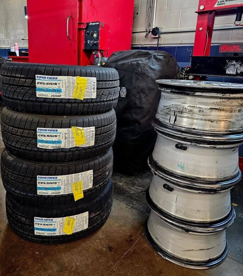 Four new tires stacked, with rims nearby in a garage setting. Tires have labels.