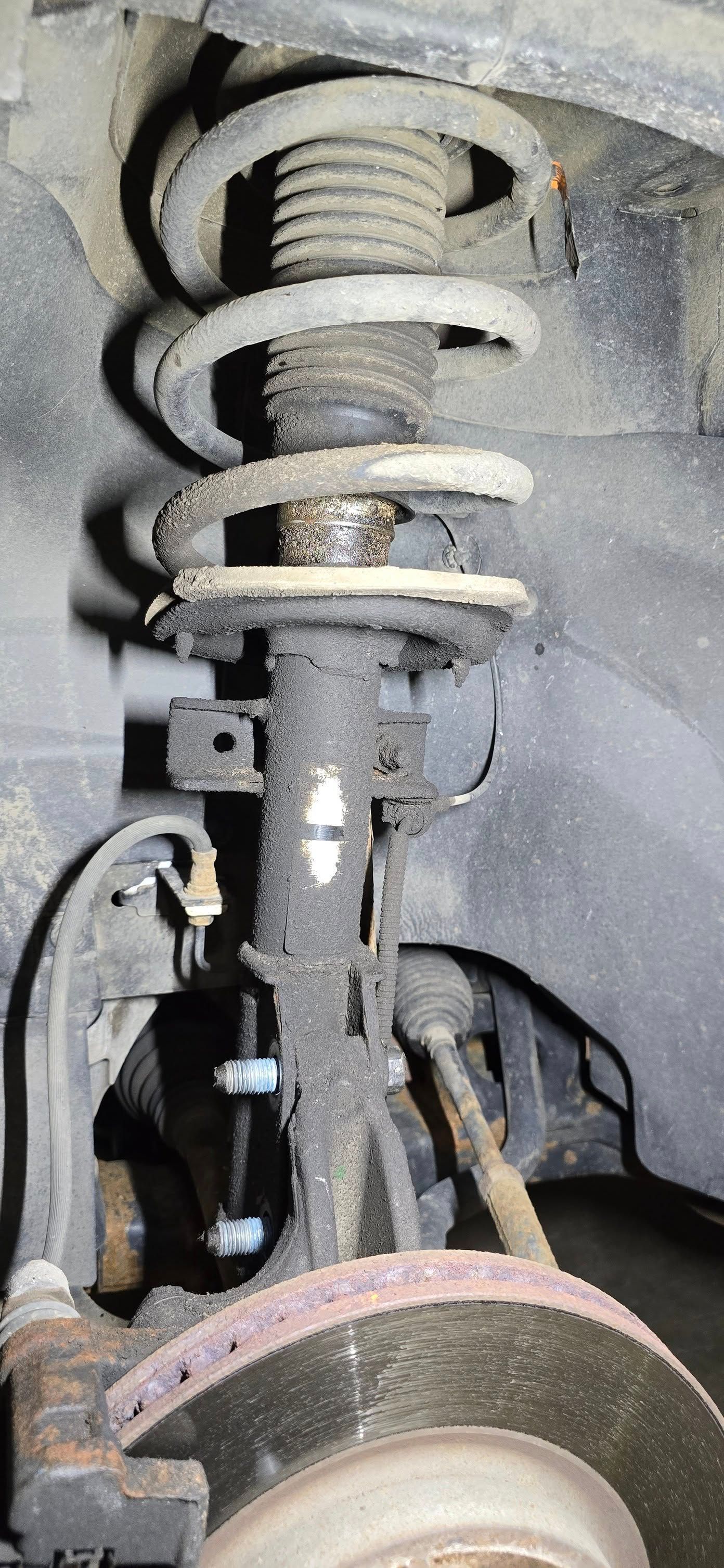 Car suspension system, likely front, shows coil spring, strut, brake rotor, and various connecting components, all covered in dirt.
