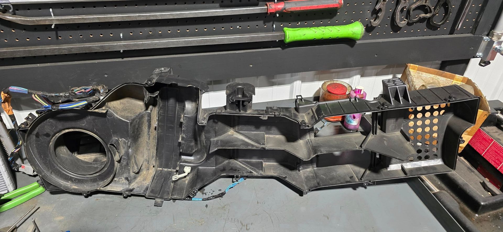 Black car air conditioning housing on a gray surface, tools on a pegboard in the background.