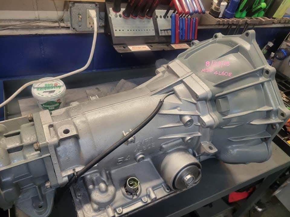 Gray automatic transmission on a workbench in a mechanic's shop. A black cable is attached.